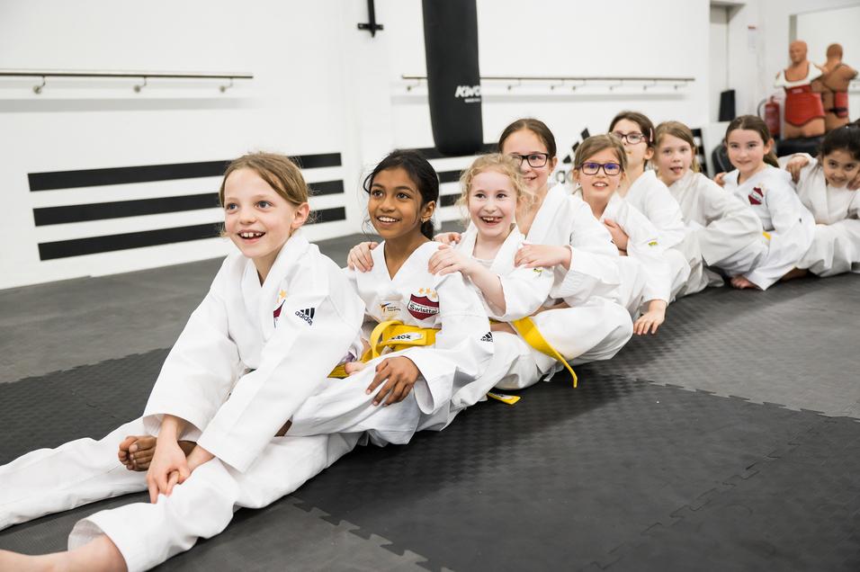 Gruppe von lächelnden Mädchen in weißen Karate-Anzügen sitzt auf einer Matte in einem Trainingsraum.