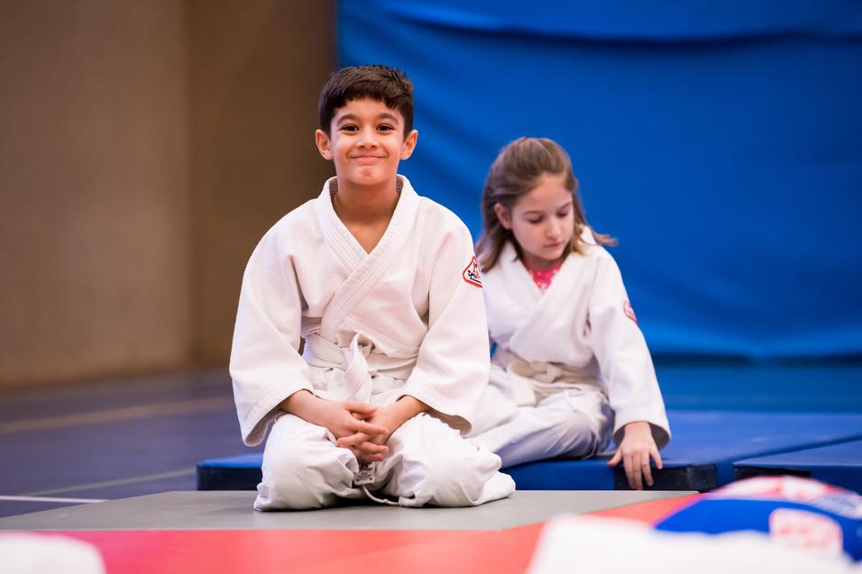 Zwei Kinder in weißen Judoanzügen sitzen auf einer Matte in einem Judoraum, der Hintergrund ist blau.