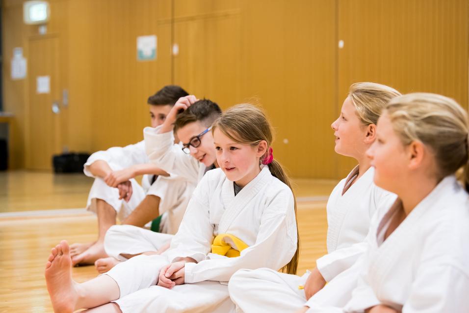 Fünf Kinder in weißen Karateanzügen sitzen in einer Trainingshalle und unterhalten sich.
