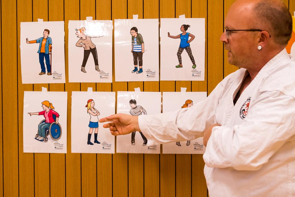 Mann im Karateanzug erklärt Poster mit verschiedenen Personen und Gesten an einer Holzwand.